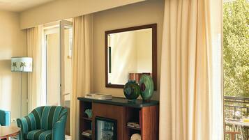 Habitación de hotel con zona de estar, estantería, espejo y vistas a espacios verdes.