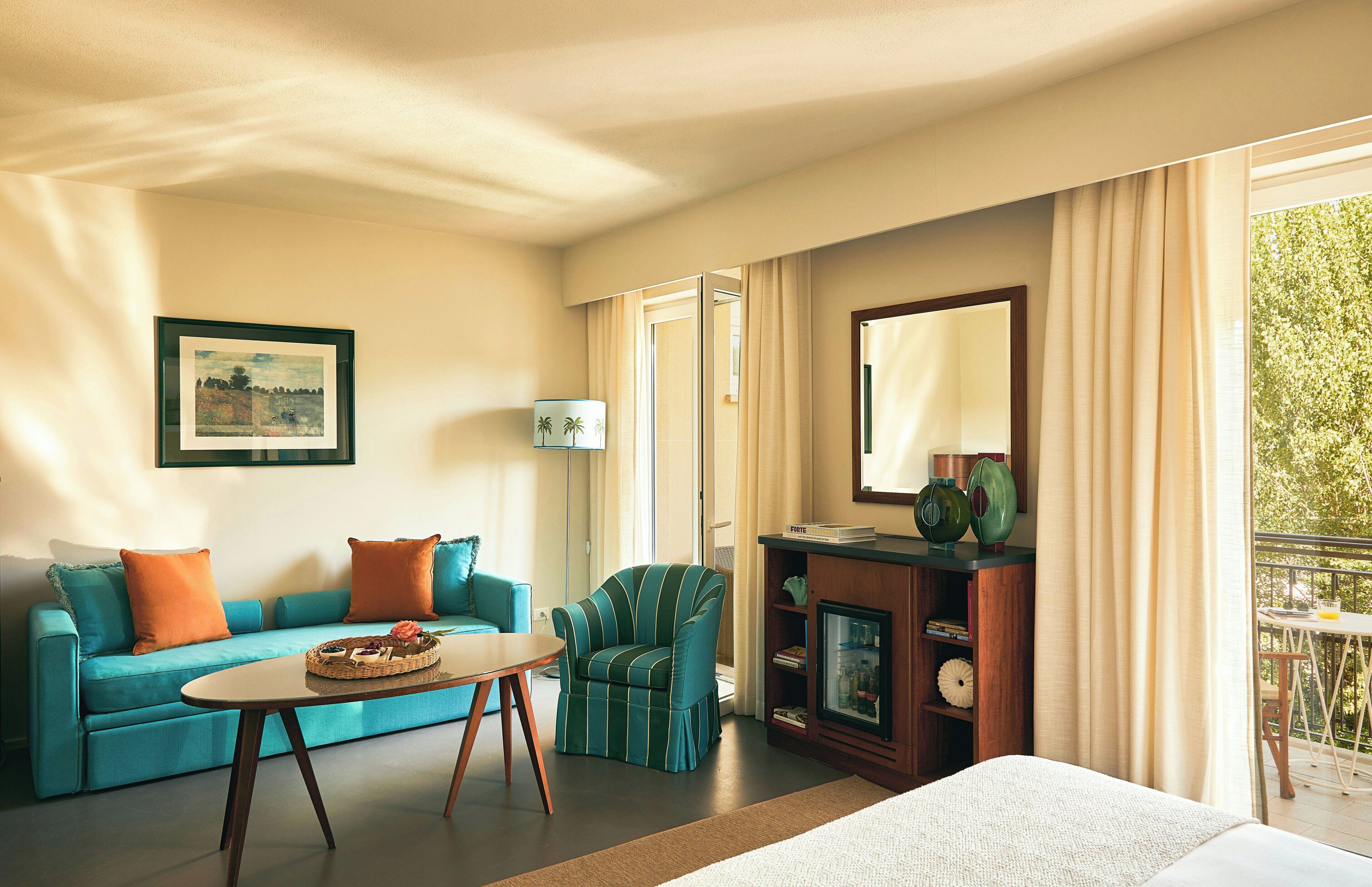 A hotel room with a bed, a sofa, a chair, a table, a bookshelf, a mirror, and a balcony with a table and chairs.