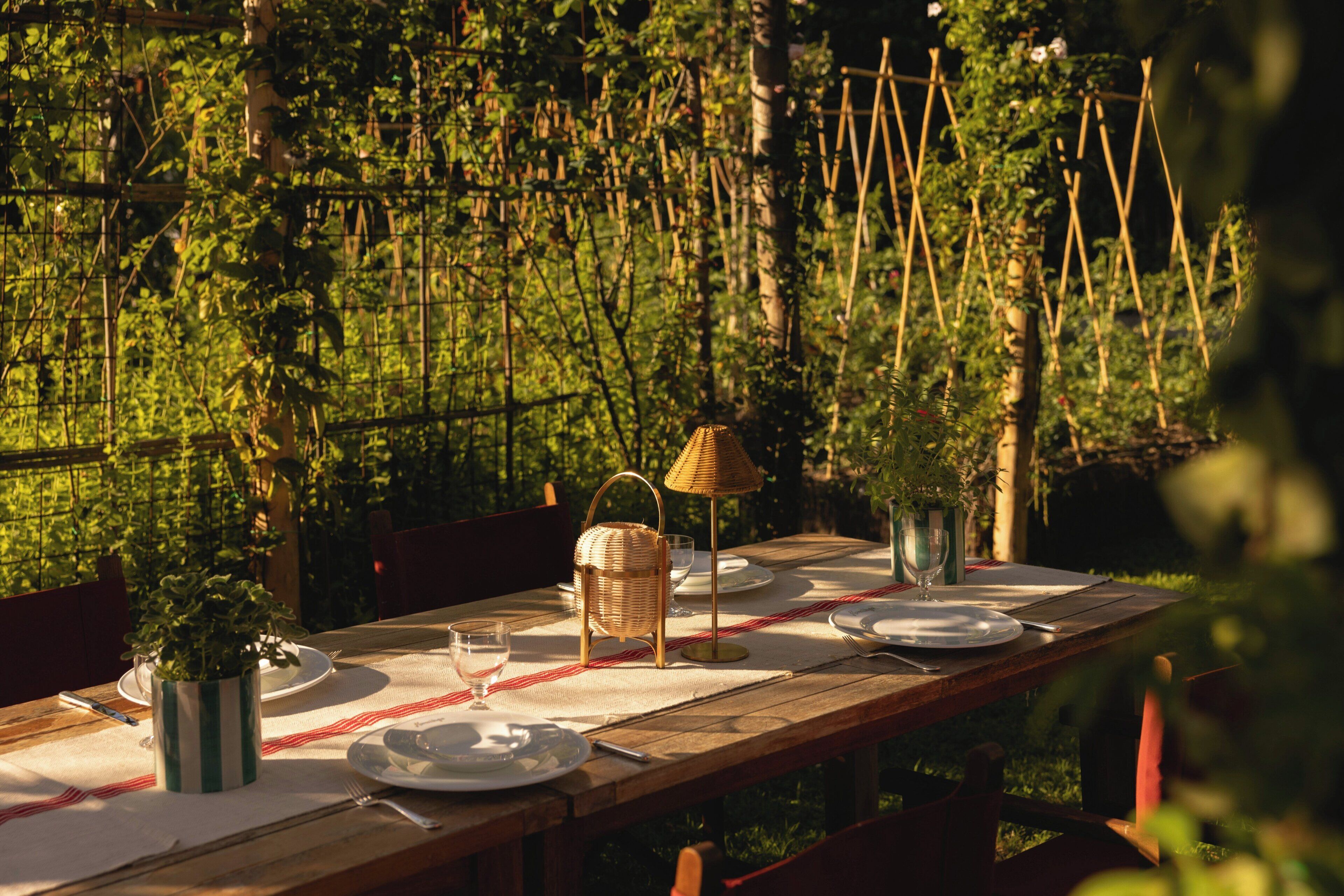 Een houten eettafel gedekt met borden, glazen en een rieten lamp, omringd door planten.
