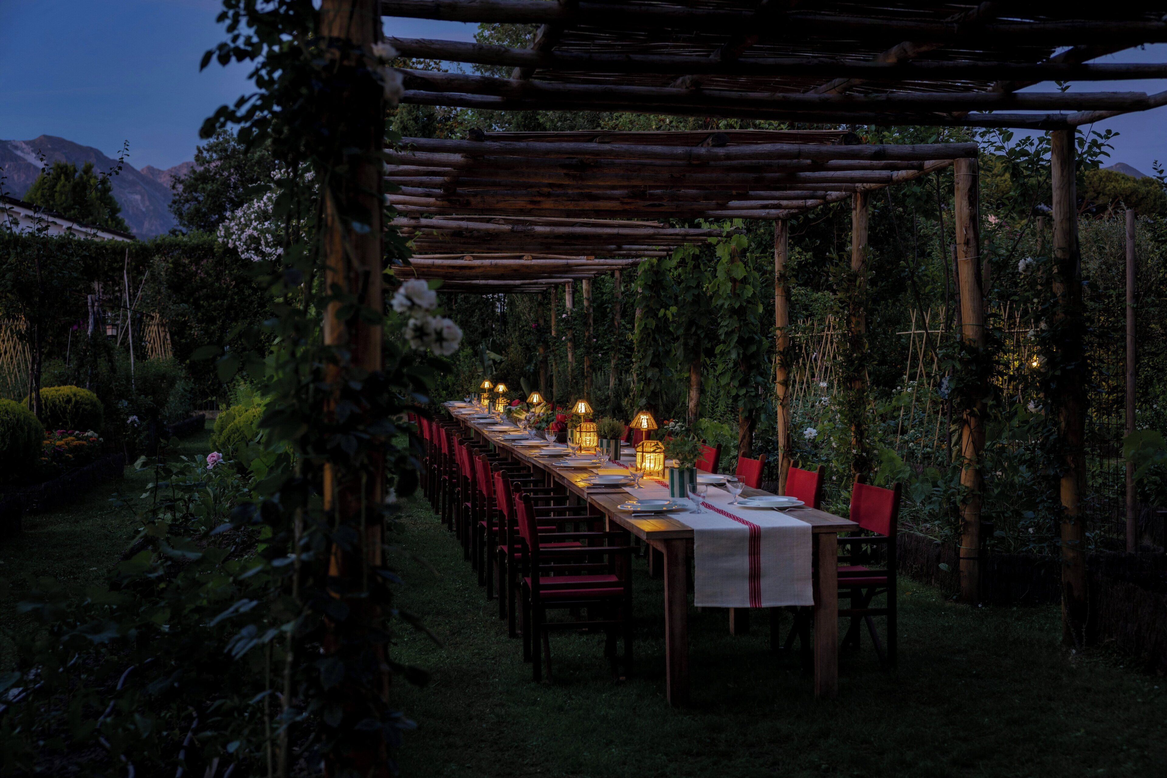 Un espace de restauration en plein air avec une longue table en bois dressée pour un repas, entourée de chaises, et abritée par une pergola en bois.
