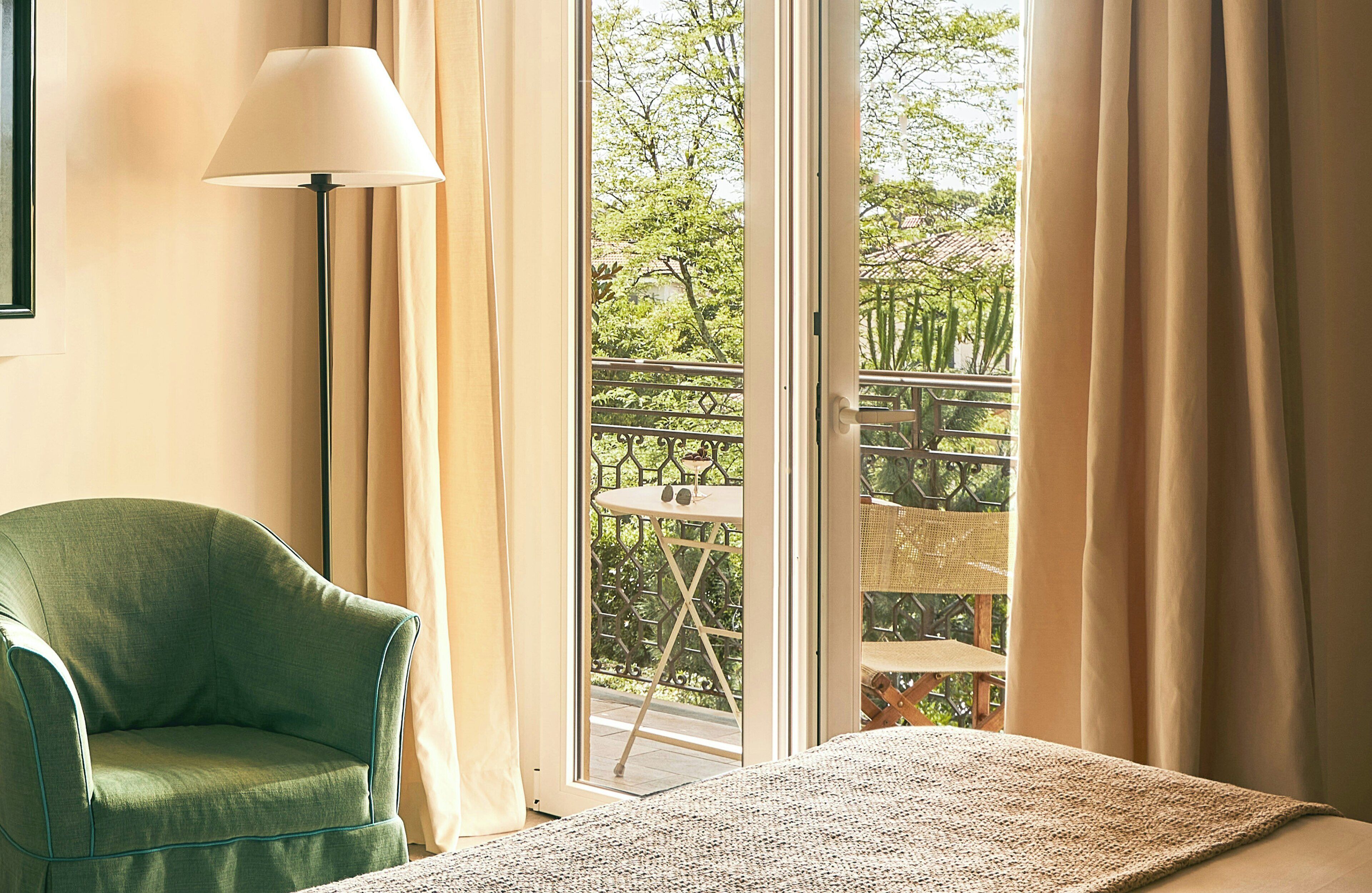 A room with a green chair, a lamp, a bed, and a view of a balcony with a table and chairs.