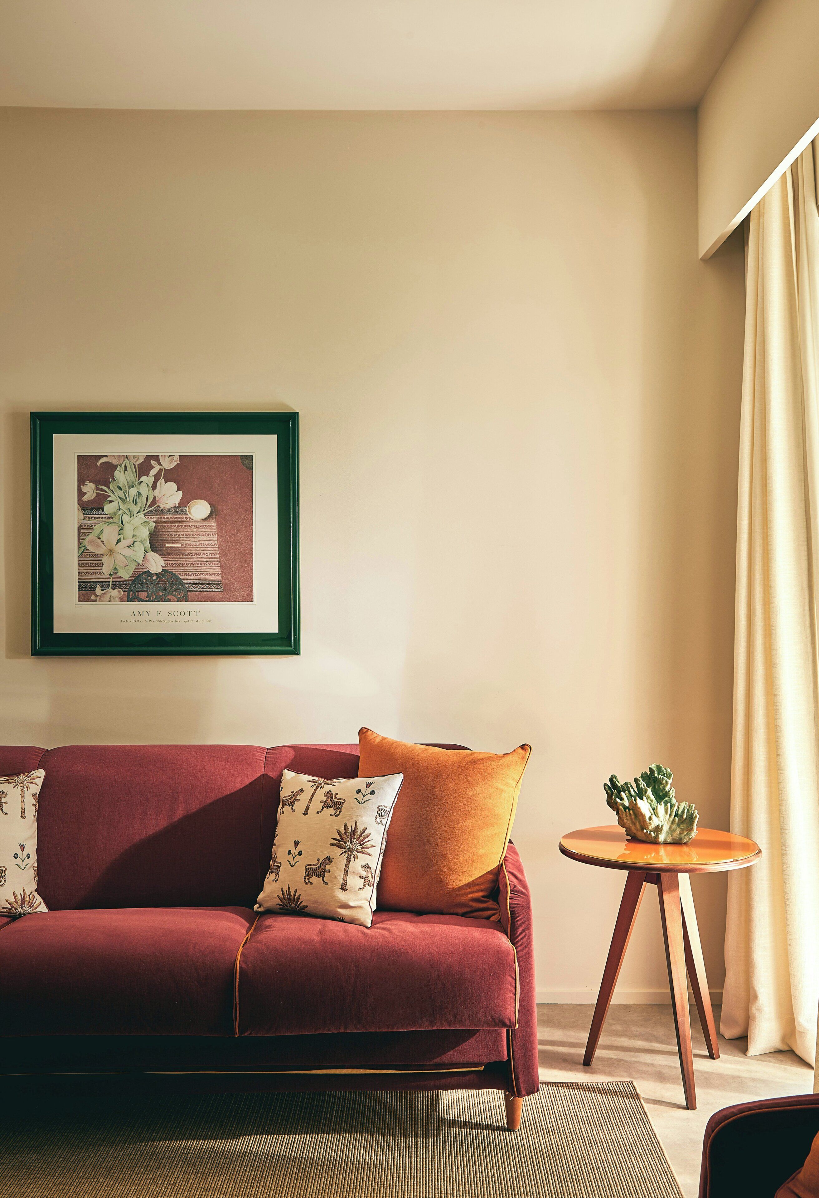 Ein Wohnzimmer mit einem roten Sofa, einem gerahmten Bild an der Wand, einem kleinen runden Tisch mit einer Pflanze und einem beigen Vorhang.