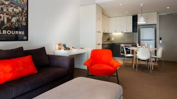 A modern living room with a dark sofa and a bright orange chair, a small dining area with a white table and chairs, and a kitchen with white cabinets and a hanging light.
