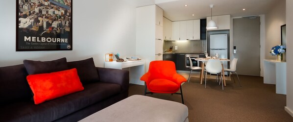Een moderne woonkamer met een donkere bank en een feloranje fauteuil, een klein eethoekje met een witte tafel en stoelen, en een keuken met witte kasten en een hanglamp.