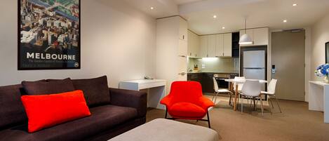 A modern hotel room with a sofa, a red chair, a dining table, and a kitchenette.