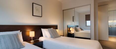 A hotel room with two beds, a nightstand with a lamp, a framed picture on the wall, and a mirror reflecting the room.