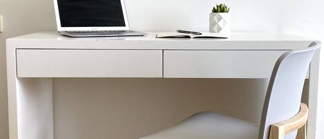 Un escritorio blanco con una computadora portátil, una pequeña planta en maceta y una silla con patas de madera.