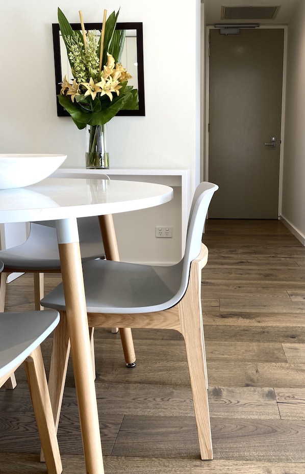 Un comedor con una mesa y sillas blancas, un jarrón con flores y suelo de madera.