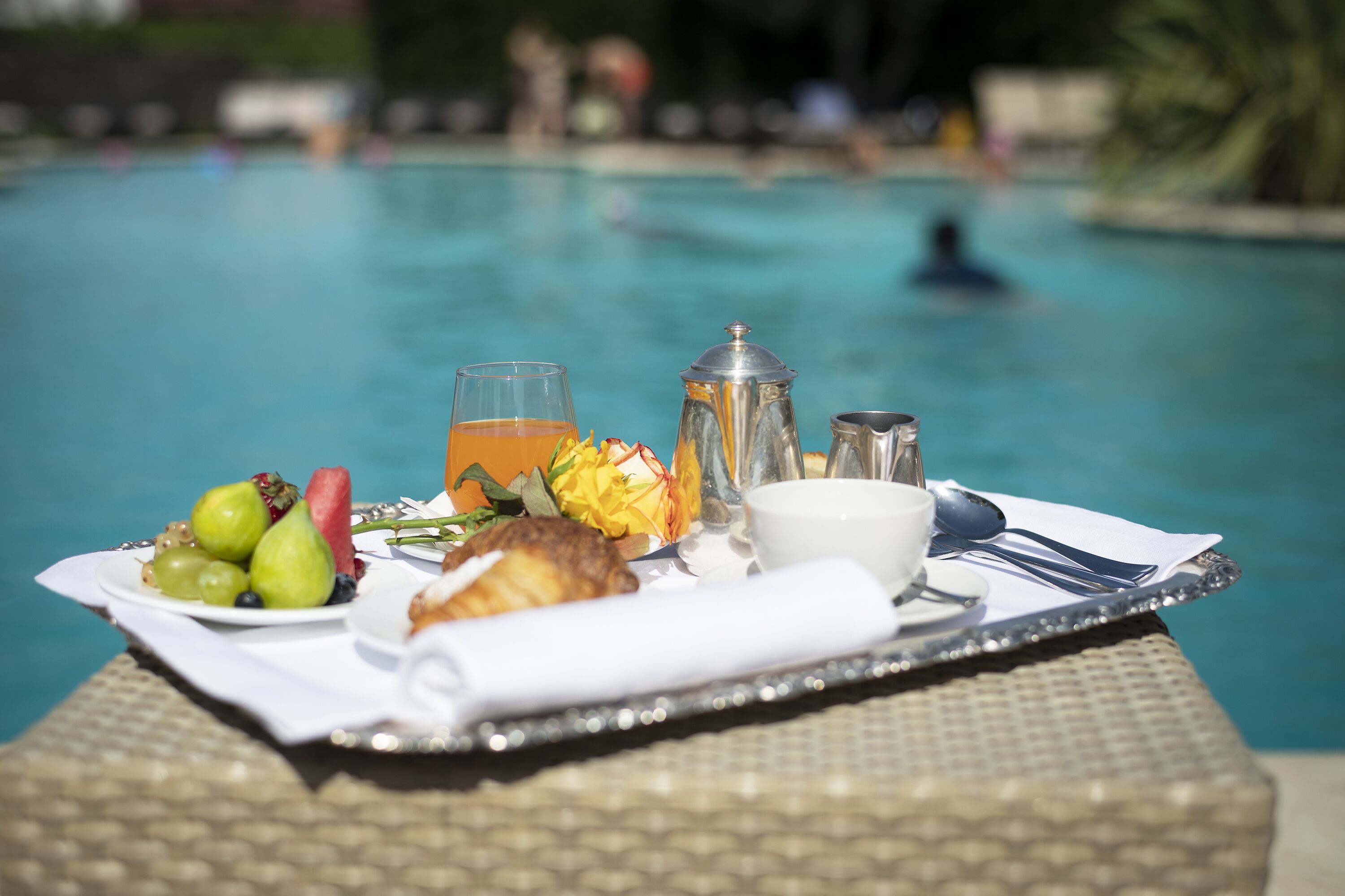 Ein Frühstücksbrett mit Früchten, einem Croissant, einem Glas Orangensaft, einer Kaffeekanne und einer Tasse Kaffee am Pool.
