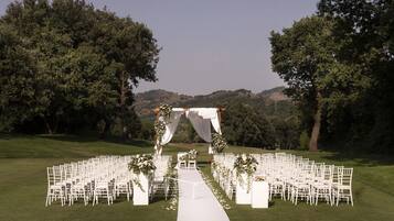 An outdoor wedding ceremony setup with white chairs arranged in rows, a white aisle, and a canopy adorned with flowers.