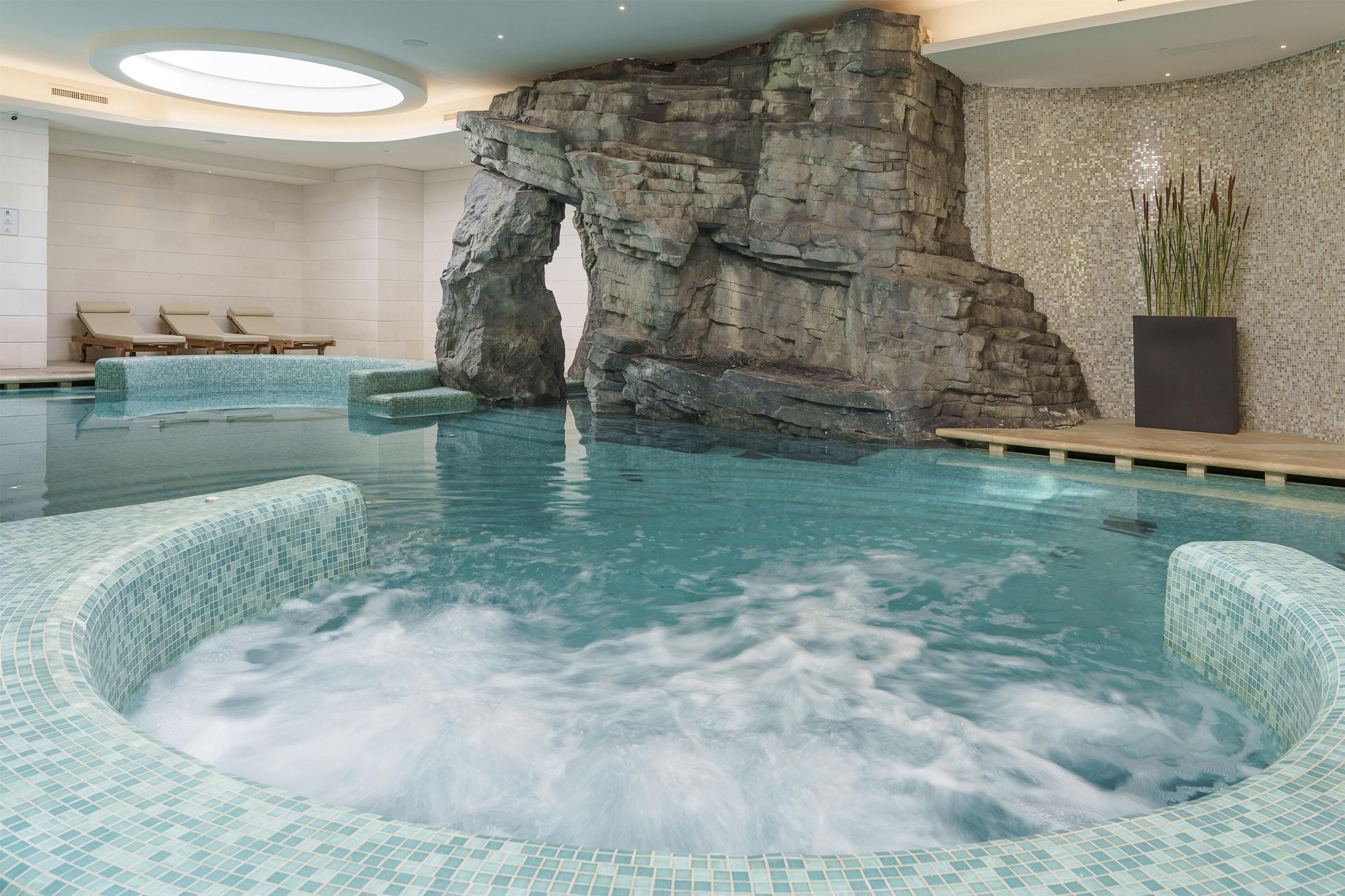 Una gran piscina interior con una formación rocosa como elemento central, sillas de descanso y una pared de azulejos.