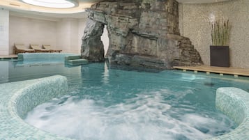 Una gran piscina interior con una formación rocosa como elemento central, sillas de descanso y una pared de azulejos.