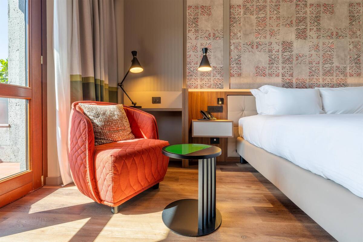 Ein Hotelzimmer mit einem Bett, einem roten Sessel, einem kleinen Tisch, einer Lampe und Blick ins Freie.
