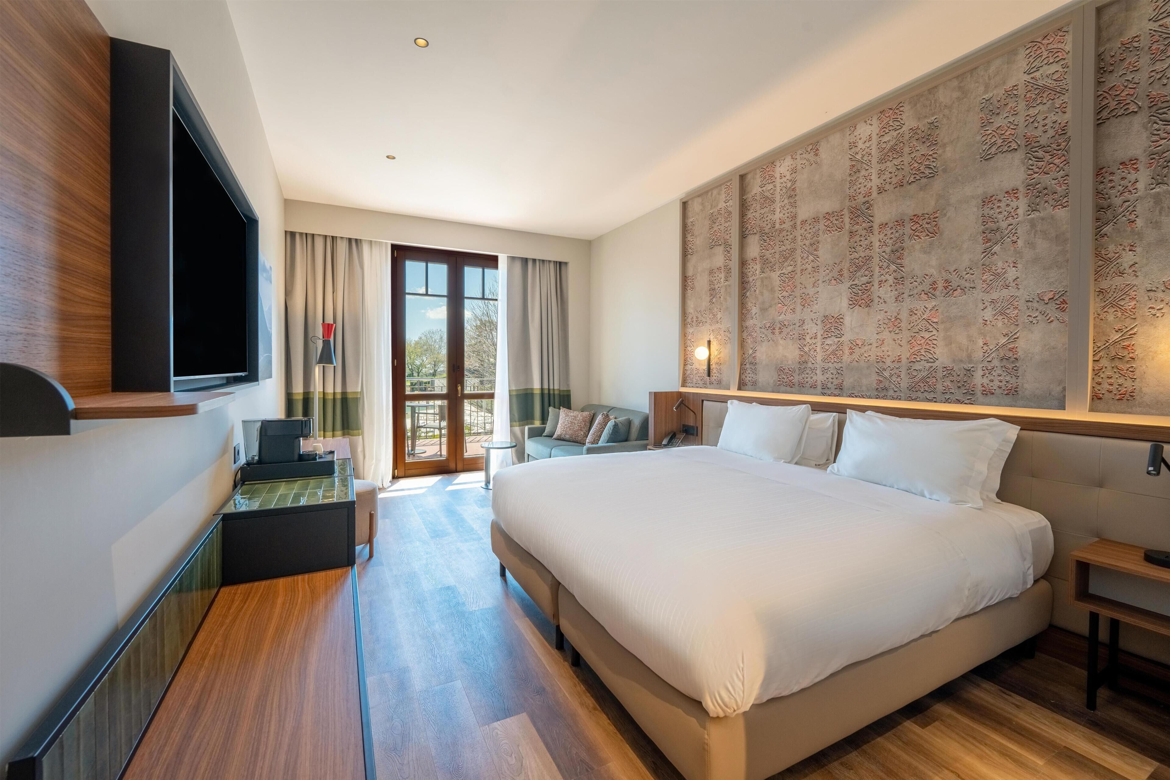 A modern hotel room with a large bed, a desk, a flat-screen TV, and a large window with curtains.