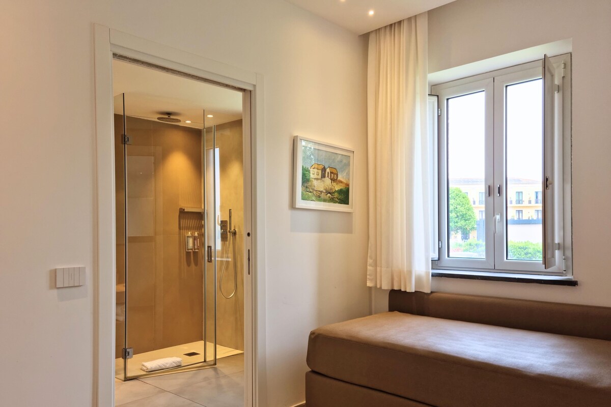 A room with a sliding glass door leading to a shower area, a sofa, a window with curtains, and a painting on the wall.