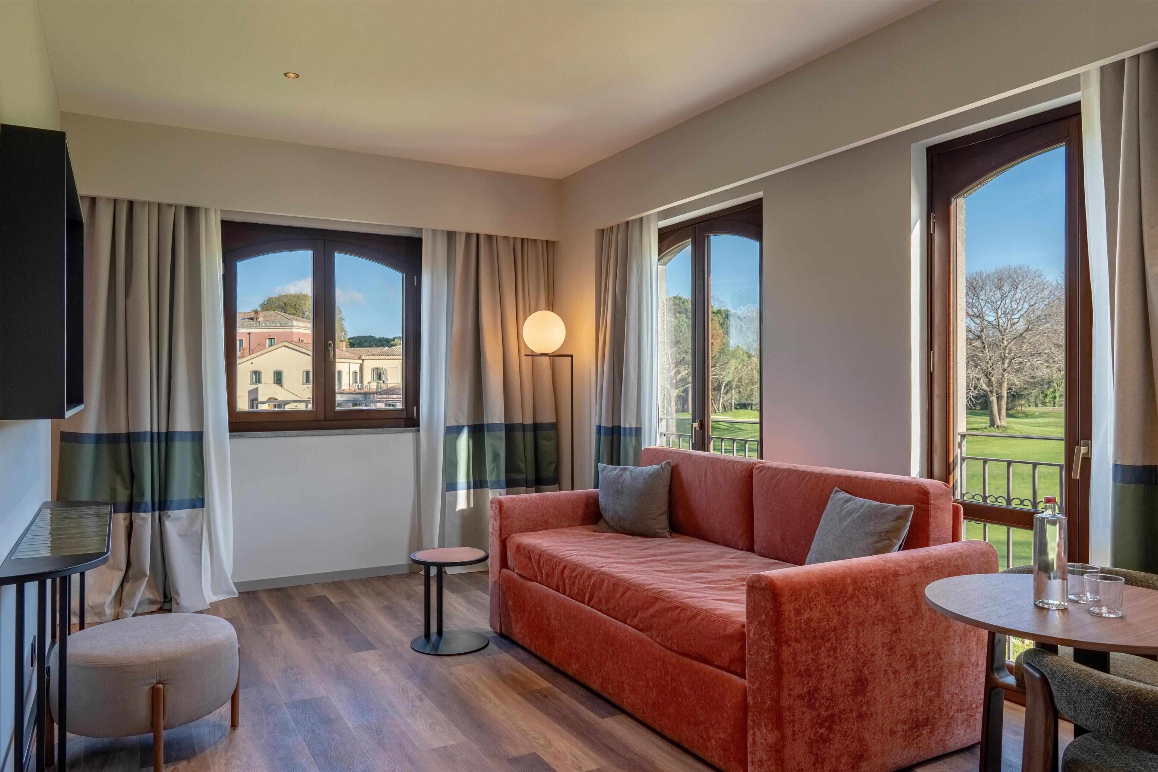 A modern living room with a sofa, a small table, a round table, and a TV. Large windows offer views of buildings and a golf course.