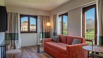 A modern living room with a sofa, a small table, a round table, and a TV. Large windows offer views of buildings and a golf course.