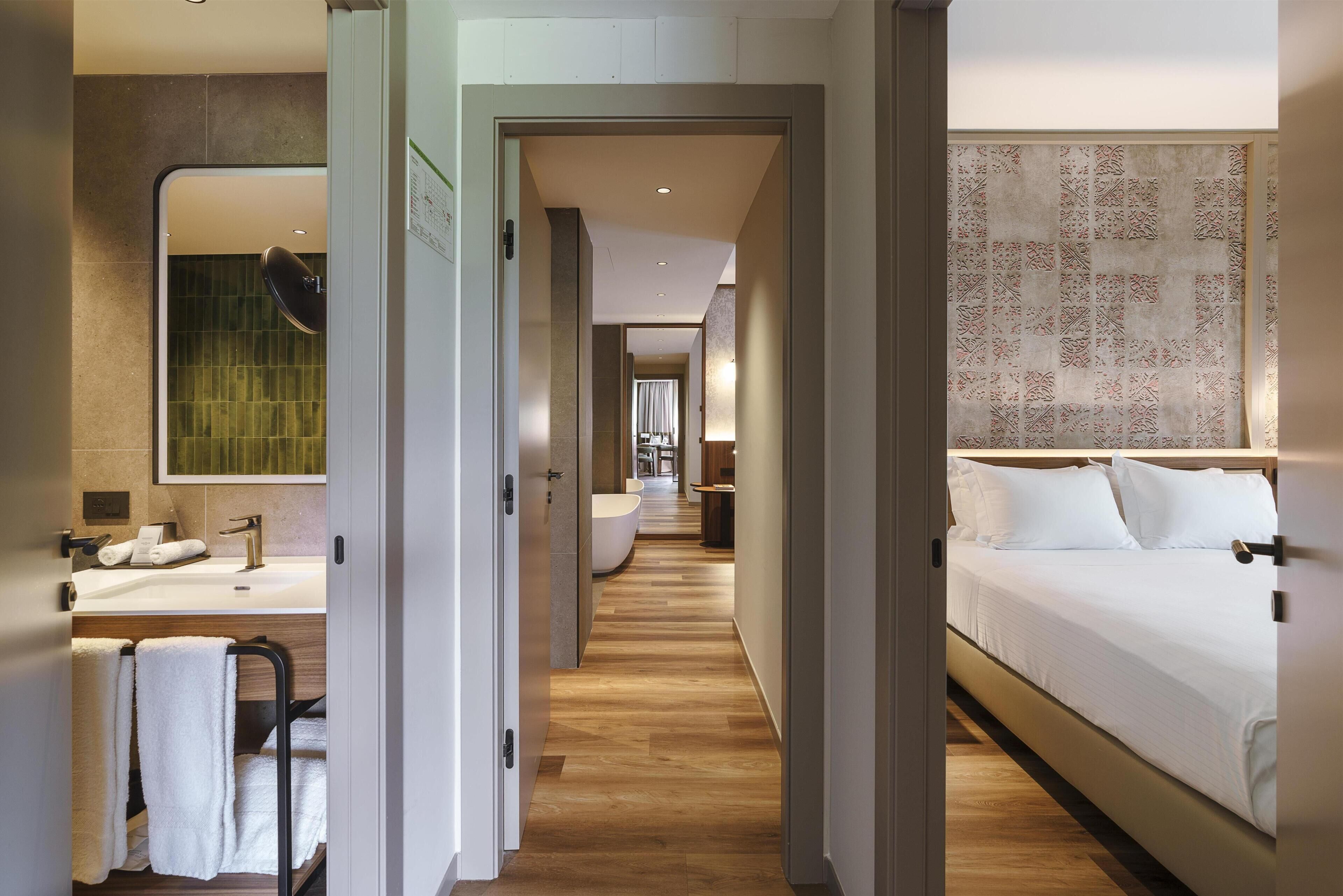 Ein Hotelzimmer mit Holzboden, einem großen Bett, einem Badezimmer mit Waschbecken und Spiegel sowie einem Gang zu einem weiteren Zimmer.