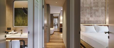 Une chambre d’hôtel avec un plancher en bois, un grand lit, une salle de bain avec un lavabo et un miroir, et un couloir menant à une autre pièce.