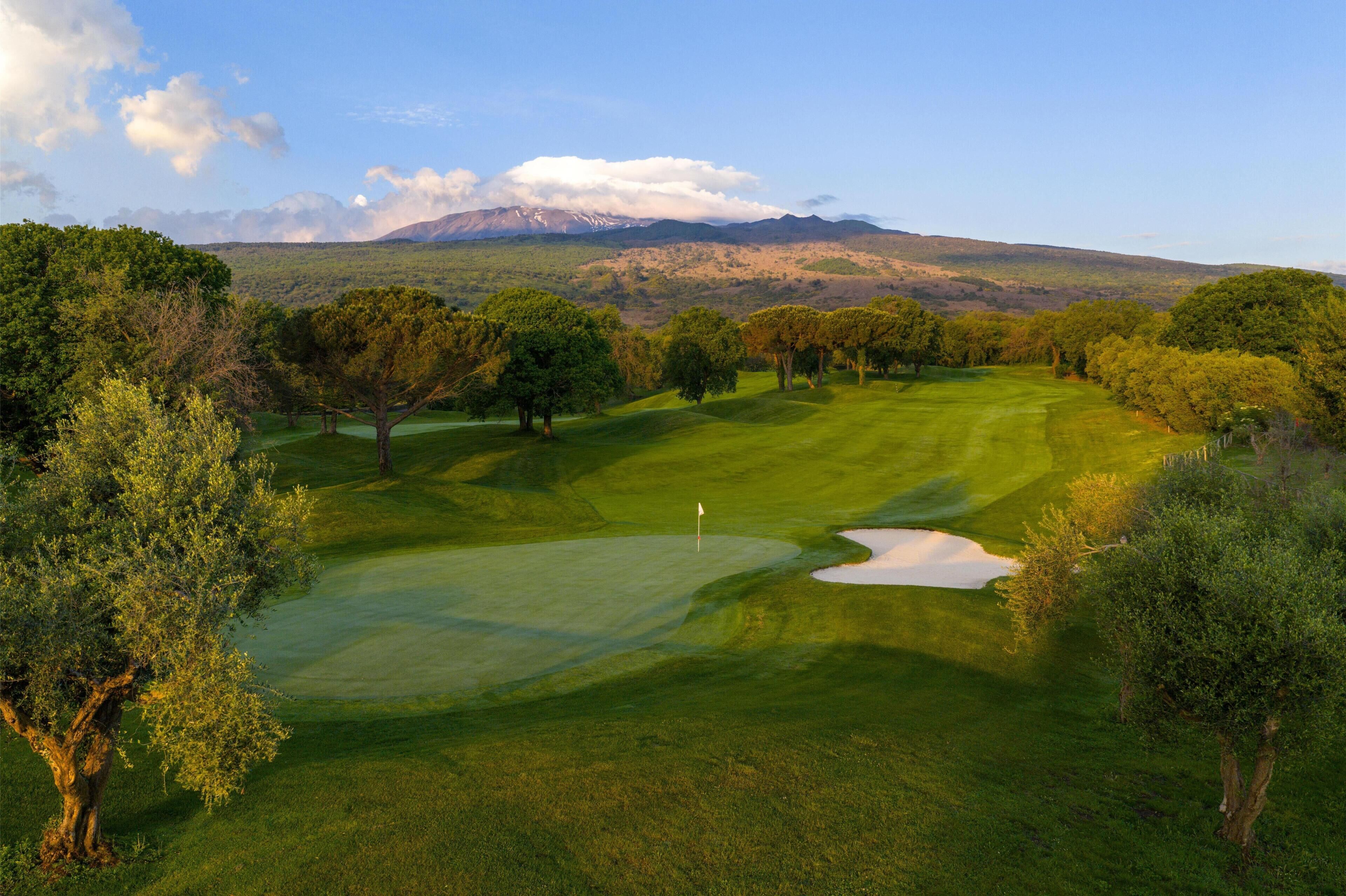 Un campo de golf con una bandera, árboles y una montaña al fondo.
