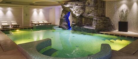 An indoor swimming pool with a rock wall, lounge chairs, and a hot tub.