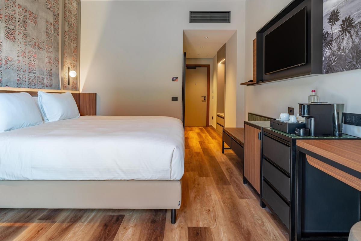 A hotel room with a bed, a desk with a coffee machine, a TV, and a door leading to another room.
