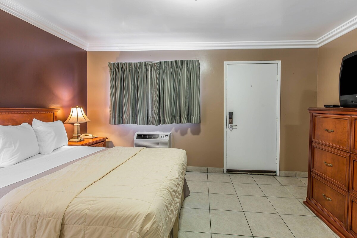 Habitación de hotel con cama, mesita de noche con lámpara, unidad de aire acondicionado, cómoda de madera y televisor.