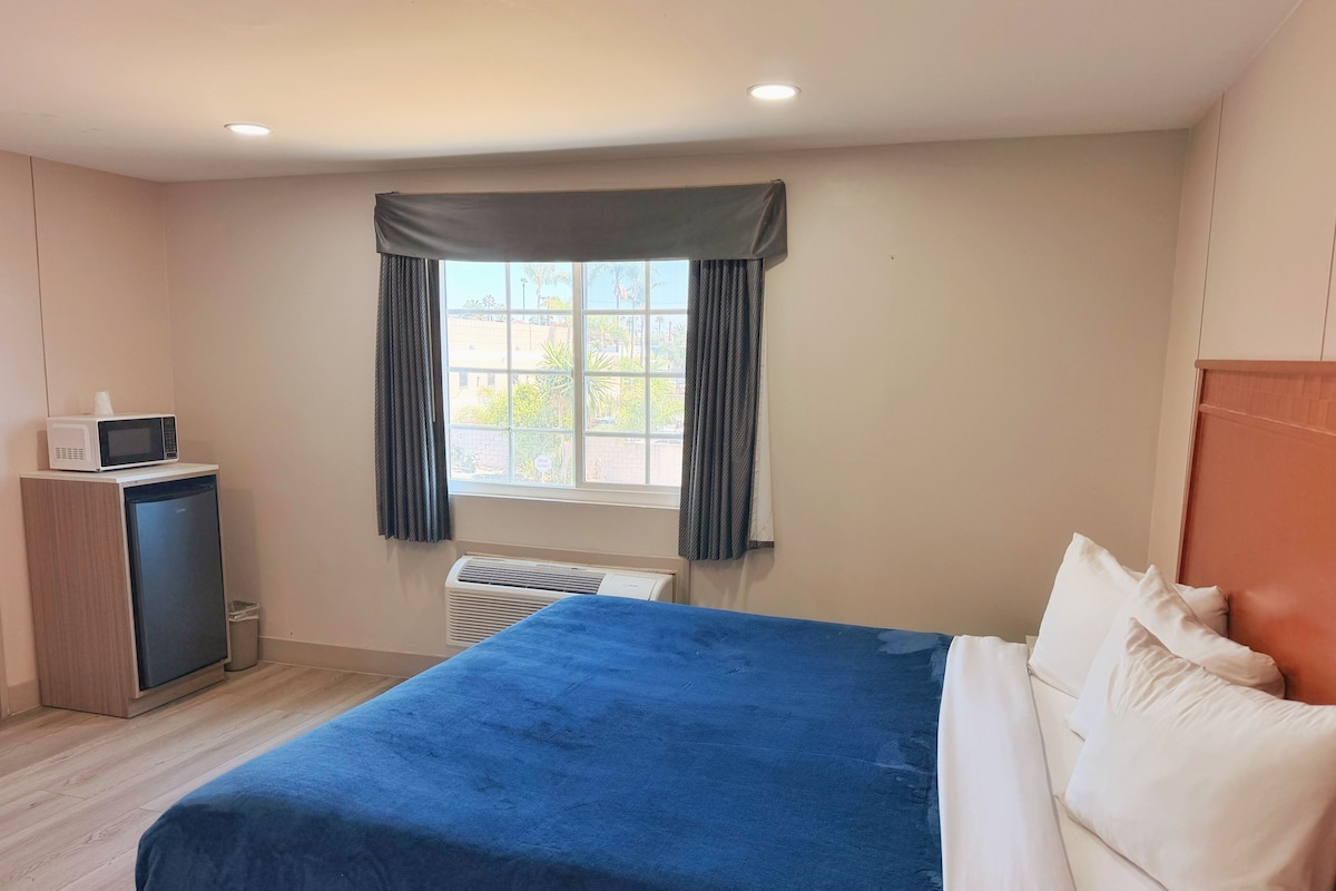 A hotel room with a bed, a small desk with a microwave, a window with curtains, and an air conditioning unit.