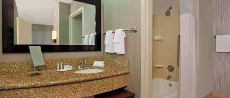 Une salle de bain avec un lavabo surmonté d’un plan de travail en granit, un miroir, une baignoire et un rideau de douche.