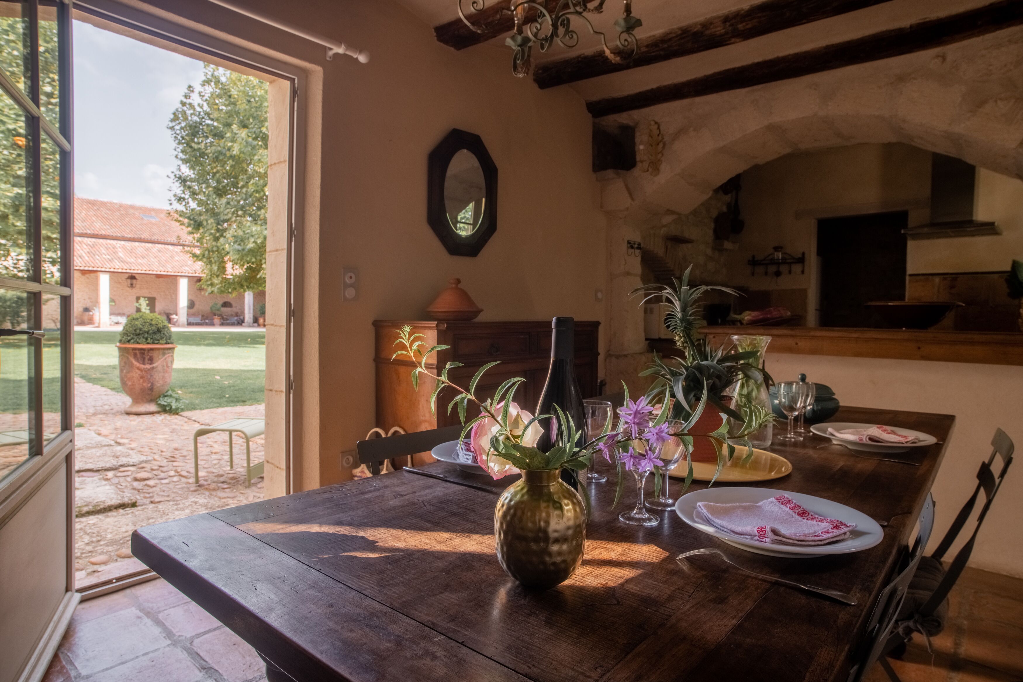 Un comedor rústico con una mesa de madera, un florero con flores y vistas a un jardín y un edificio.