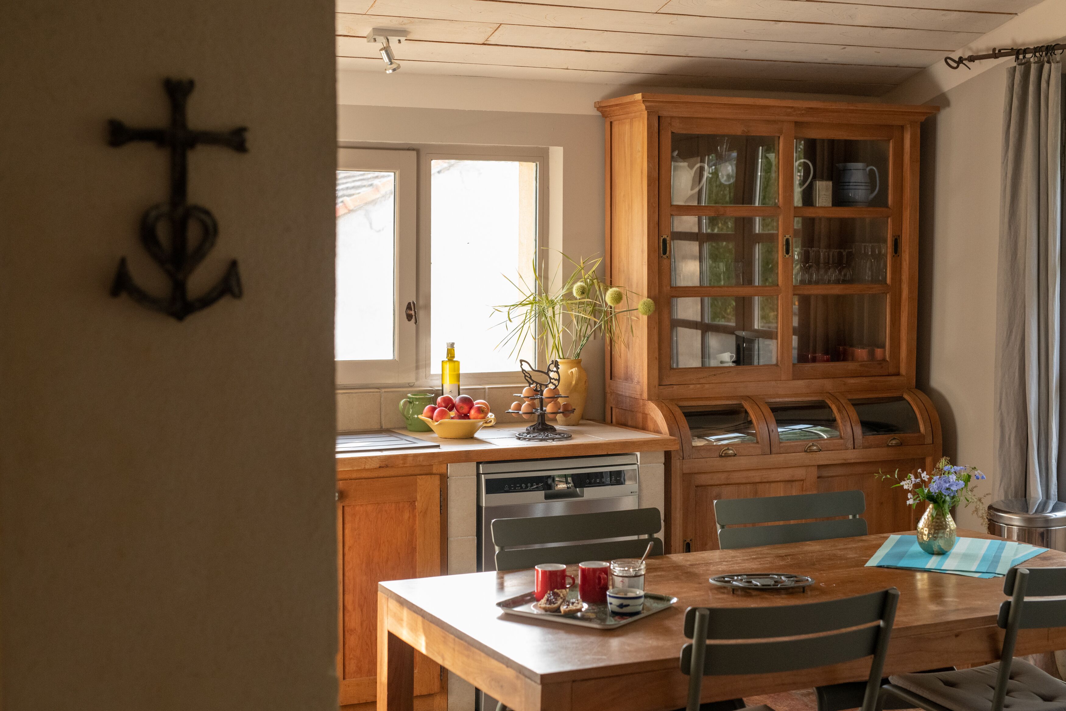 Ein Holzespeisetisch mit Stühlen, ein Küchenbereich mit einem Holzkasten und ein Fenster mit Vorhängen.