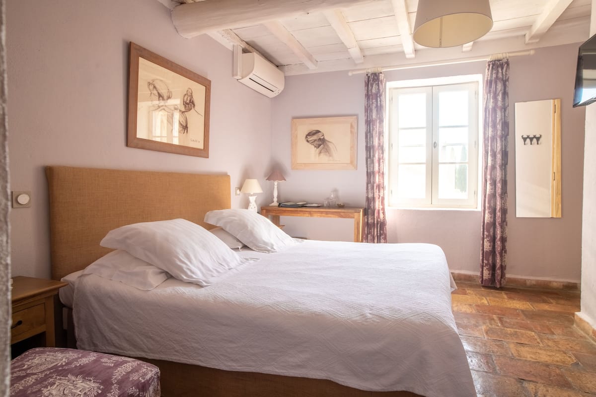 Un dormitorio con cama, mesita de noche, escritorio de madera, silla, lámpara y ventana con cortinas.