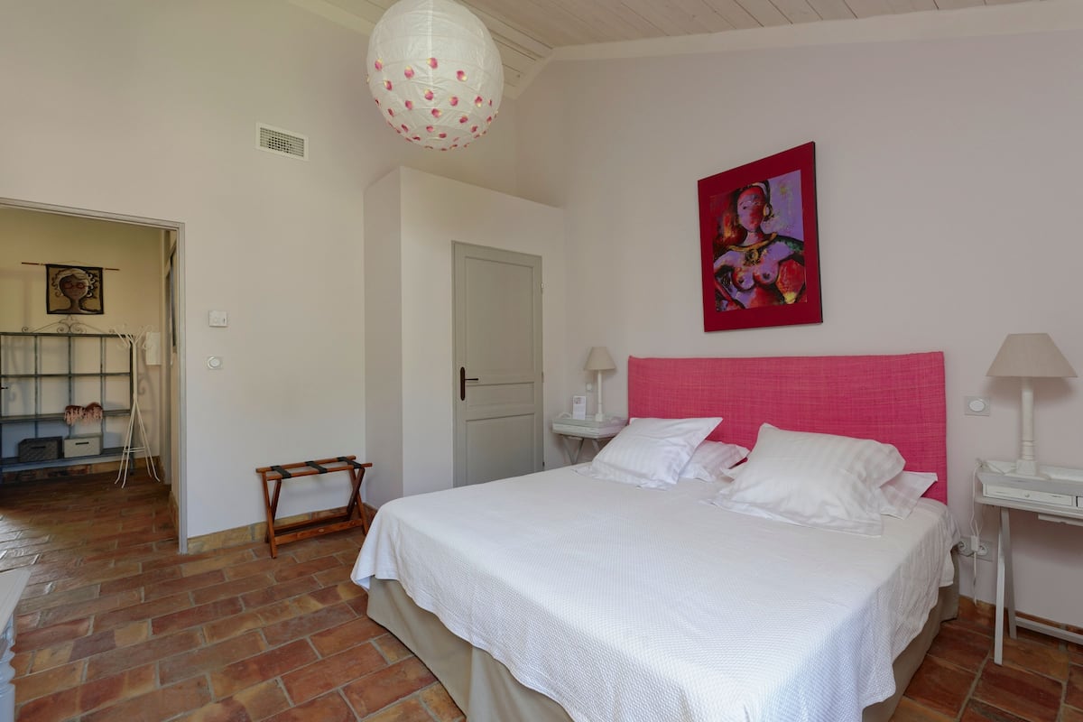 A bedroom with a large bed, a white headboard, a bedside table with a lamp, and a door leading to another room.