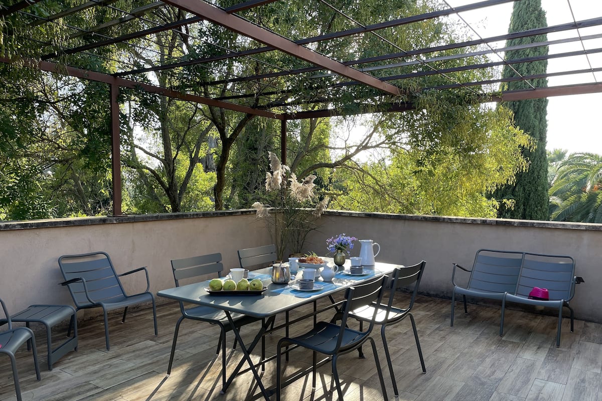 Un espacio exterior techado con una mesa dispuesta para dos personas, sillas y un banco alrededor.