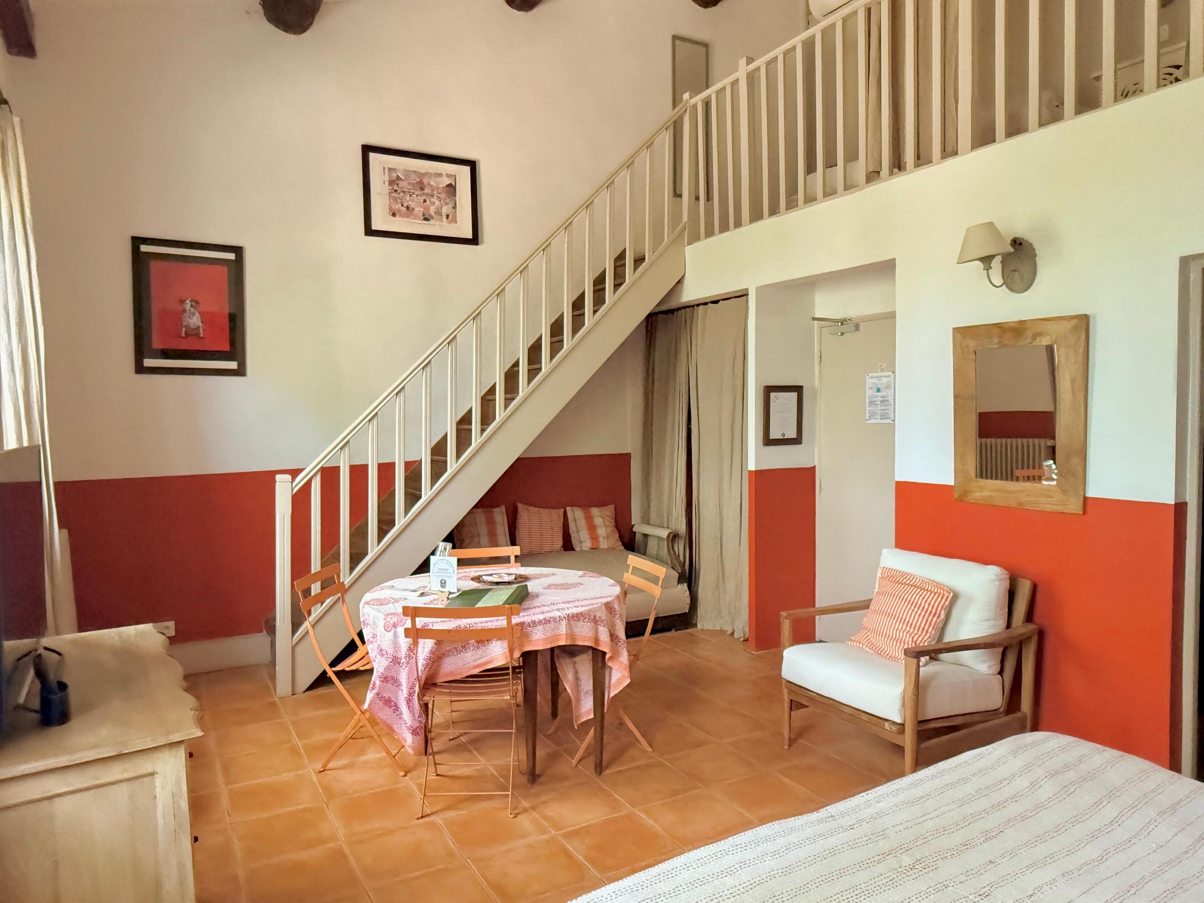 A room with a dining table, chairs, a sofa, a staircase, and a bed.