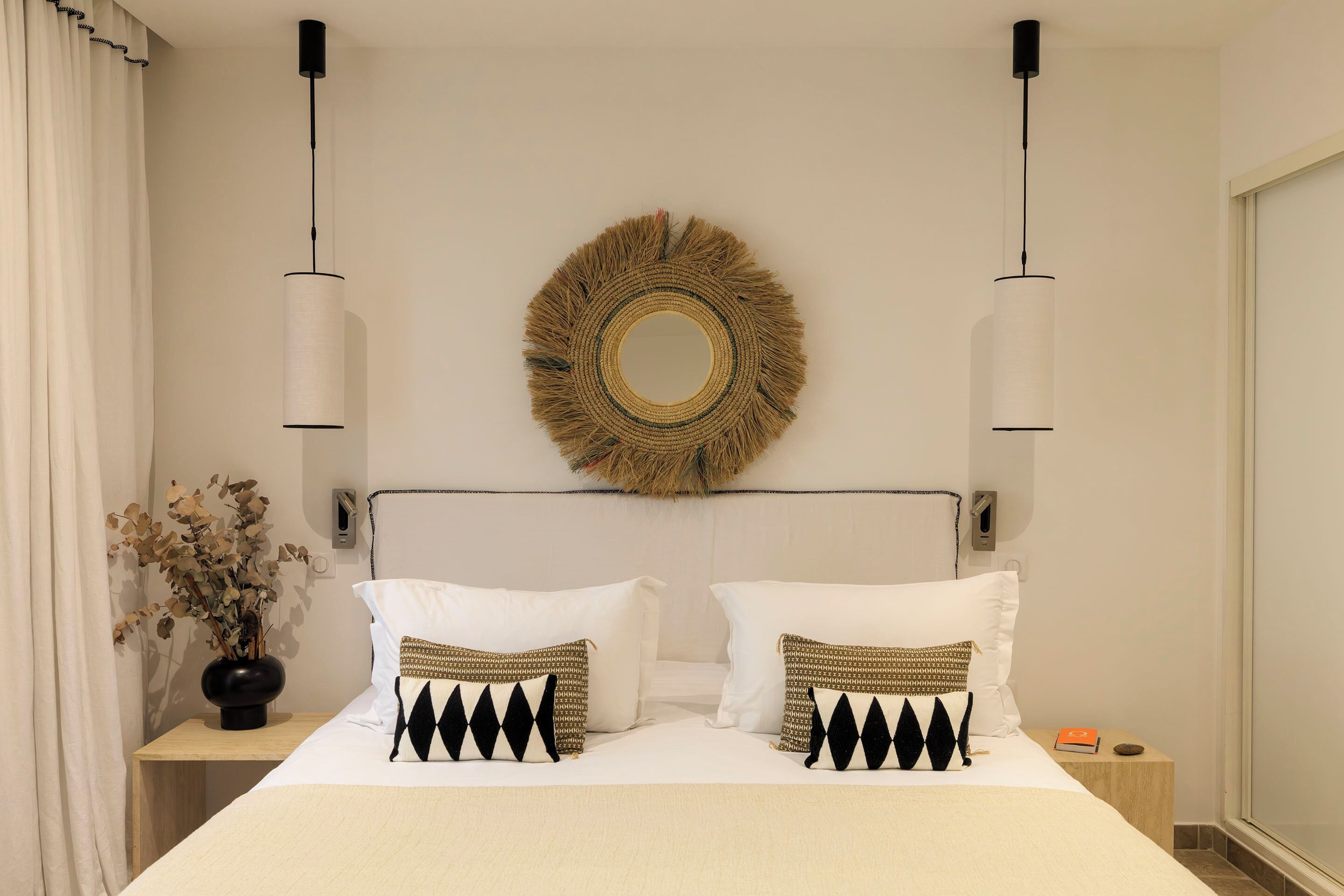 Una cama bien hecha con dos almohadas, un espejo montado en la pared y dos lámparas colgantes.