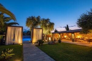 Un patio exterior bien iluminado con un sendero de piedra que lleva a una piscina, rodeado de exuberante vegetación y un edificio de piedra con asientos al aire libre.