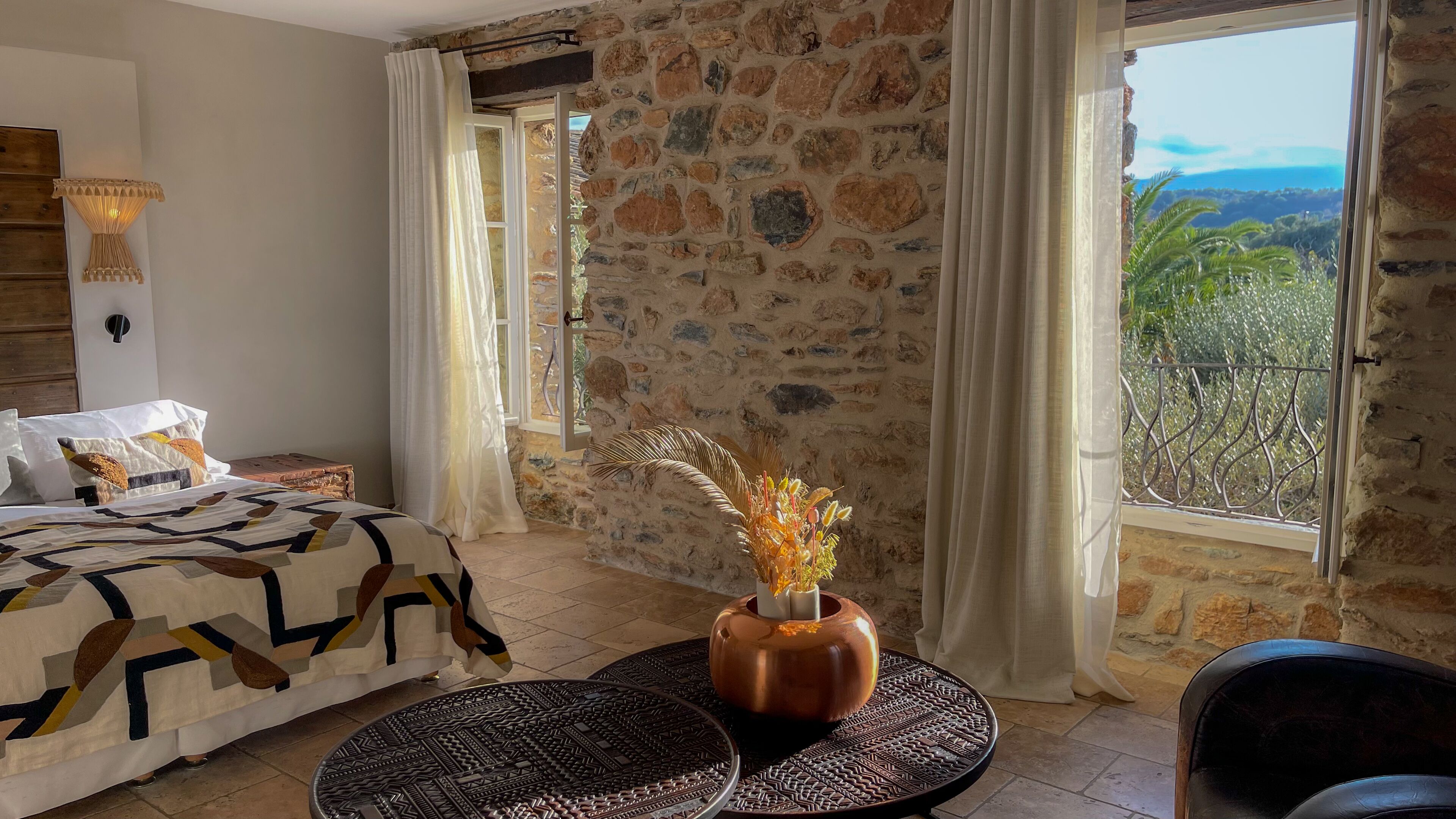 A bedroom with a stone wall, a bed with patterned bedding, a vase with dried flowers, and a view of greenery through a window.