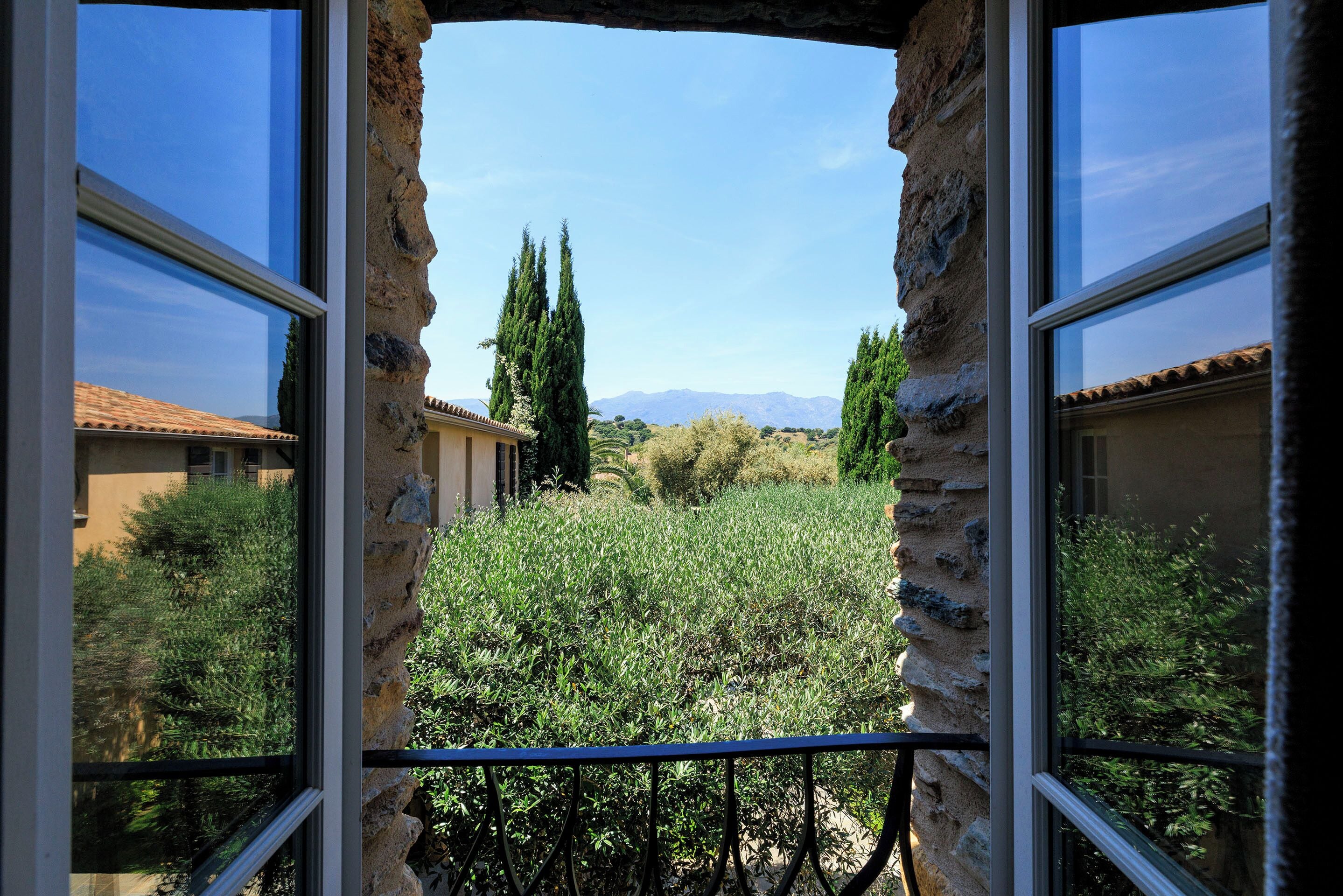 Une vue à travers une arche en pierre surmontée d’une rambarde métallique, qui mène à un jardin où poussent différentes espèces de plantes, avec une chaîne de montagnes visible au loin.