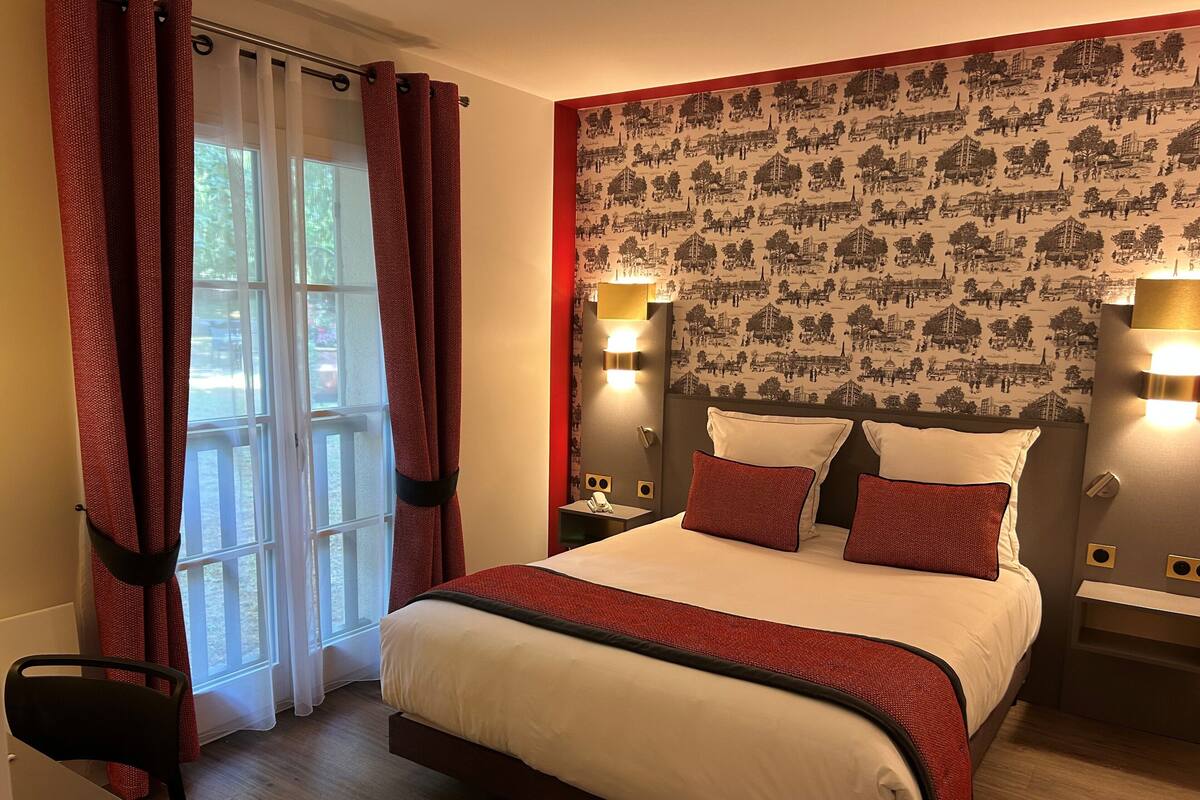 A hotel room with a bed, curtains, a nightstand, a chair, and a wall with a patterned wallpaper.