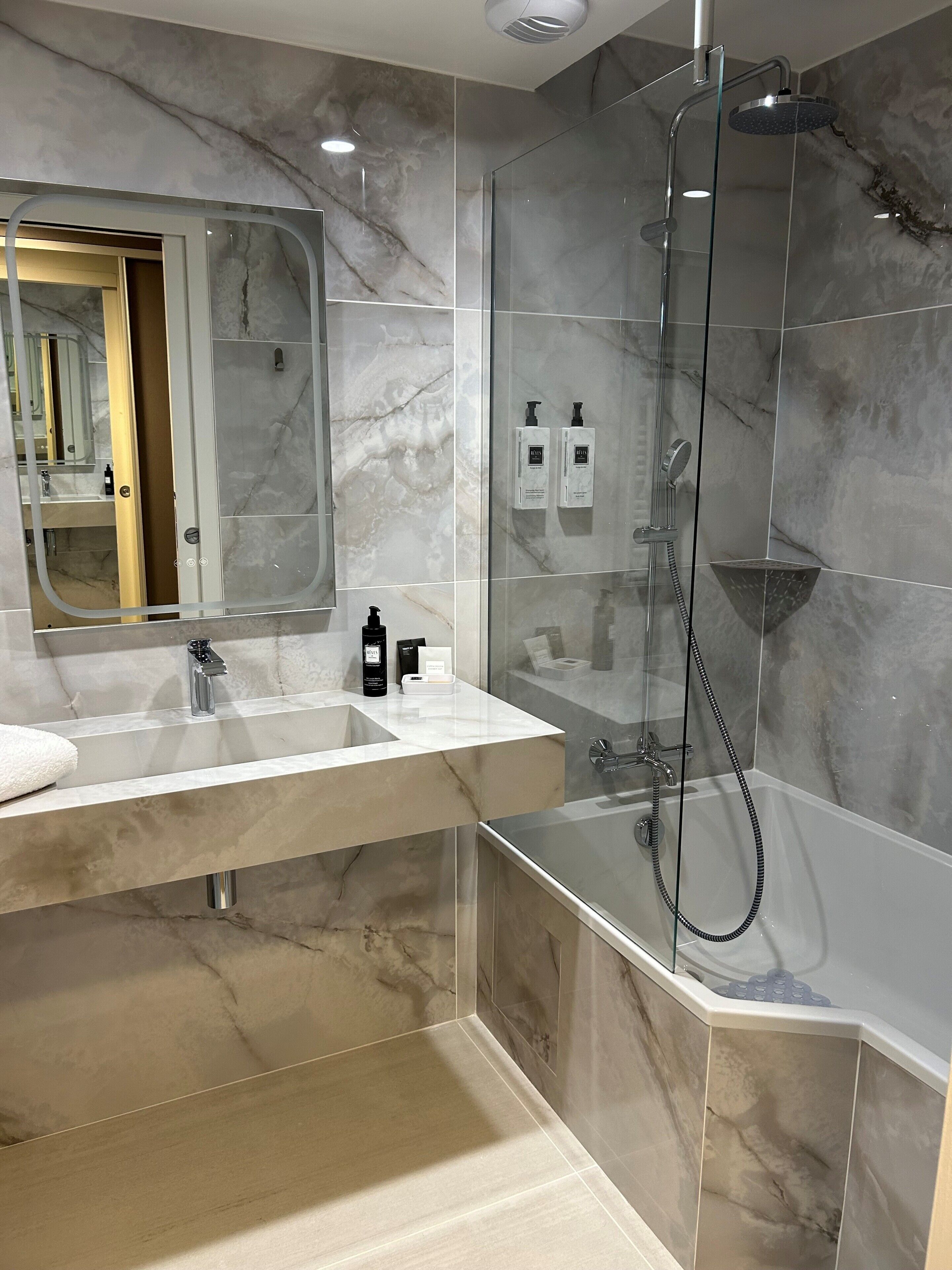 Un baño moderno con ducha envuelta en vidrio, tocador con un espejo grande y paredes de mármol.