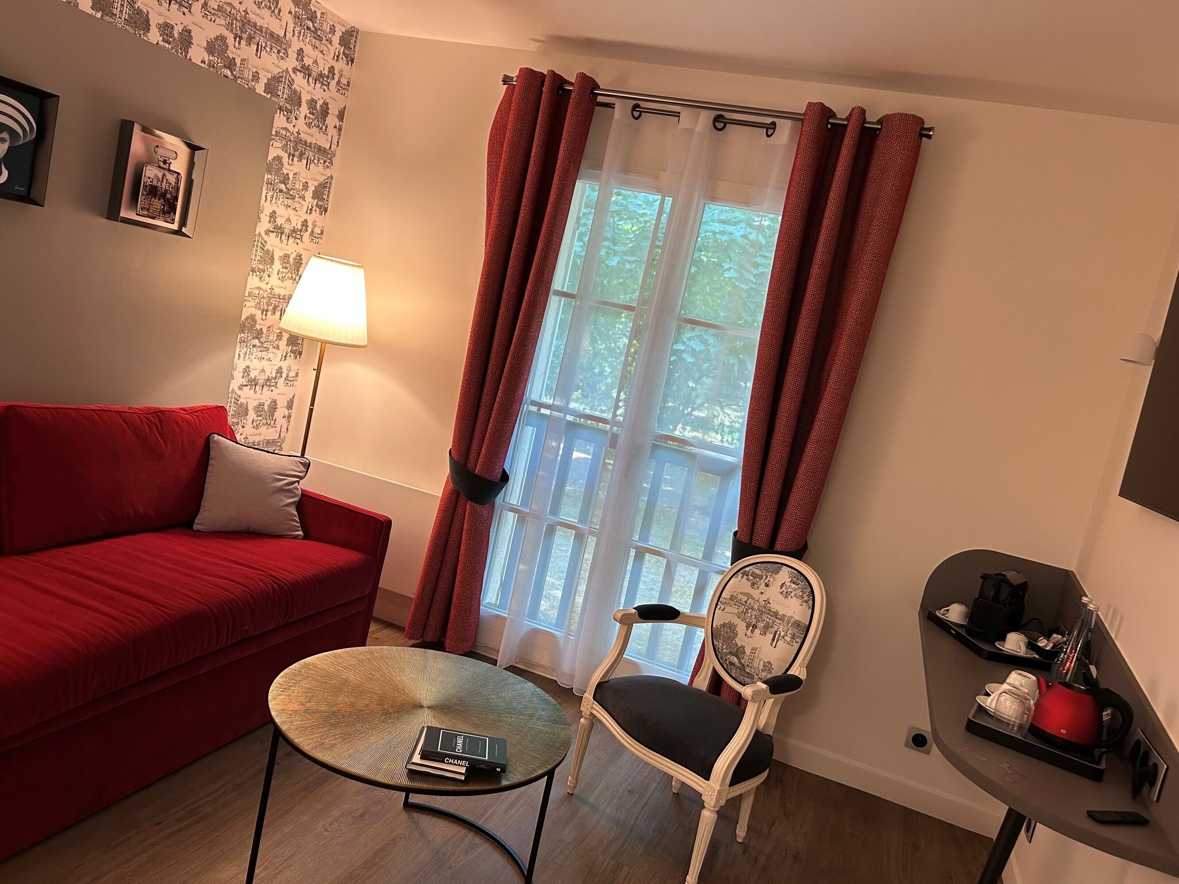 A red sofa, a small round table with a book, a chair with a patterned cushion, a window with red curtains, and a lamp.