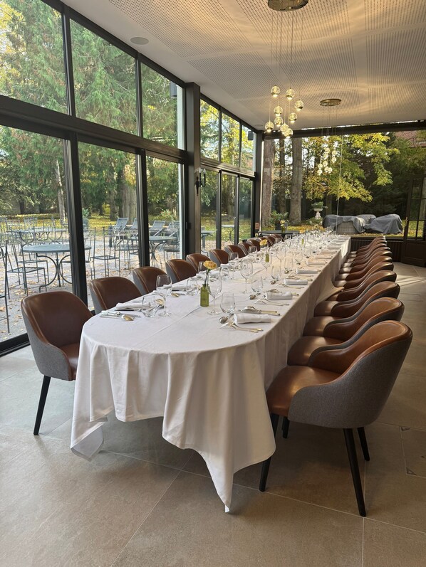 A long dining table set for a meal with chairs, large windows, and a view of trees.
