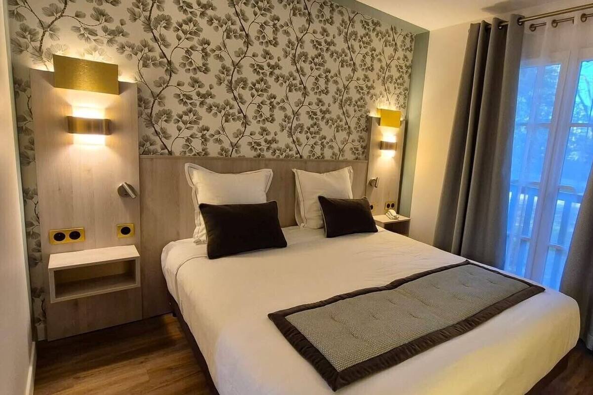 A hotel room with a bed, bedside tables, a wall-mounted light, a window with curtains, and a view of trees.
