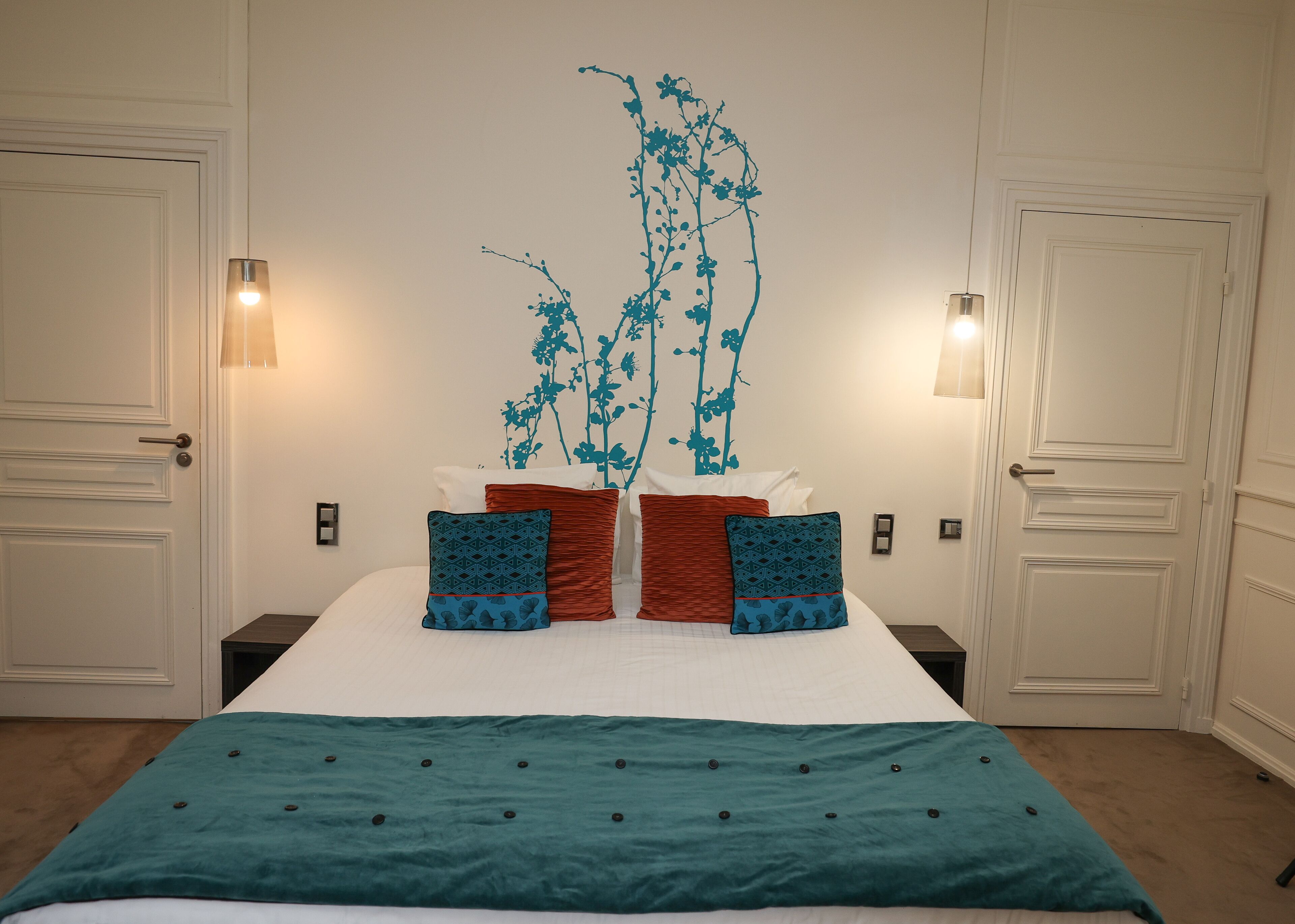 Una cama bien tendida con una colcha color turquesa y dos almohadones decorativos, flanqueada por dos lámparas de pared y una obra de arte en la pared.