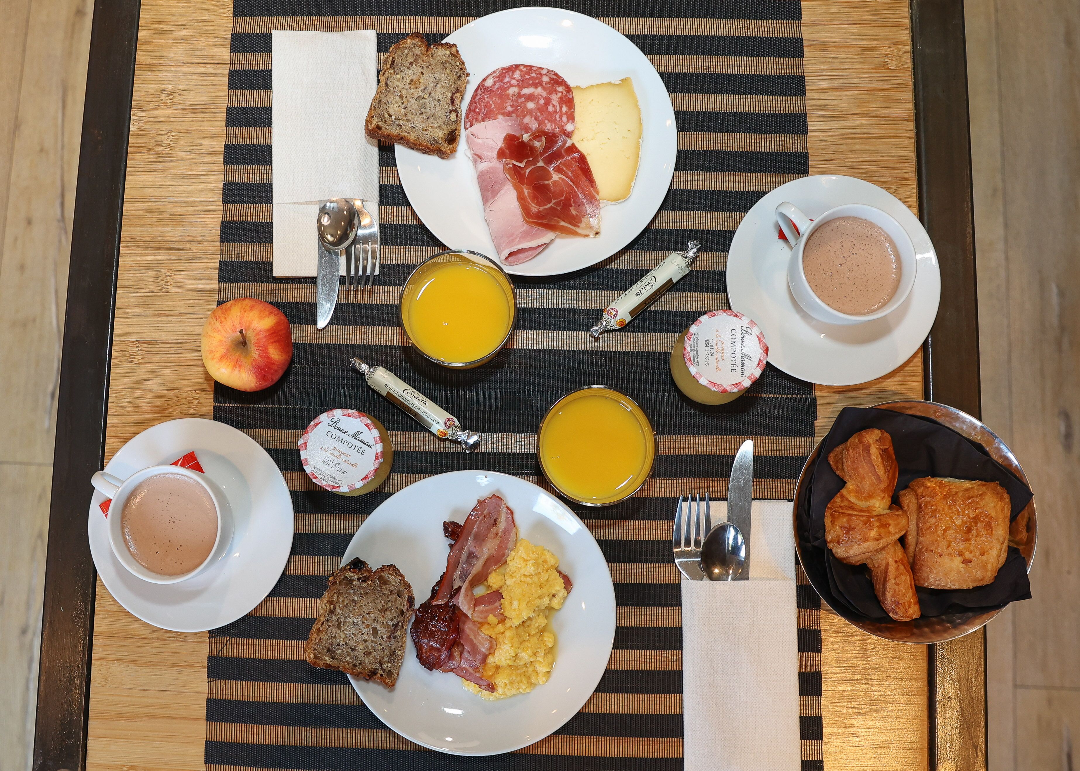 Ett frukostbord med tallrikar fyllda med ägg, bacon, ost och bröd, samt koppar med kaffe och juice.