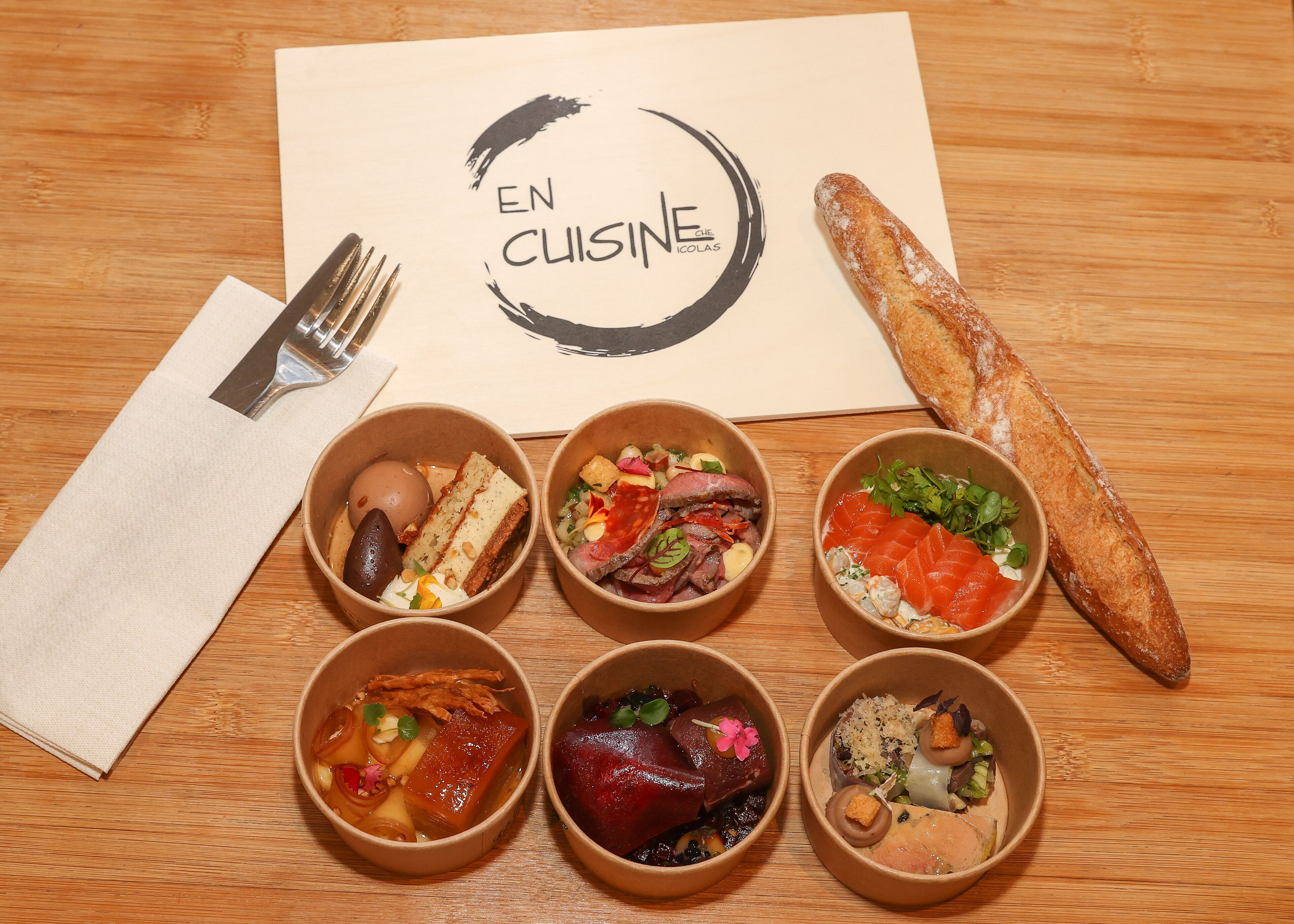 Una mesa de madera con seis fuentes de comida, una baguette y una servilleta.