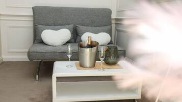 A grey sofa with heart-shaped cushions, a white coffee table with a champagne bottle and glasses, and a decorative vase.