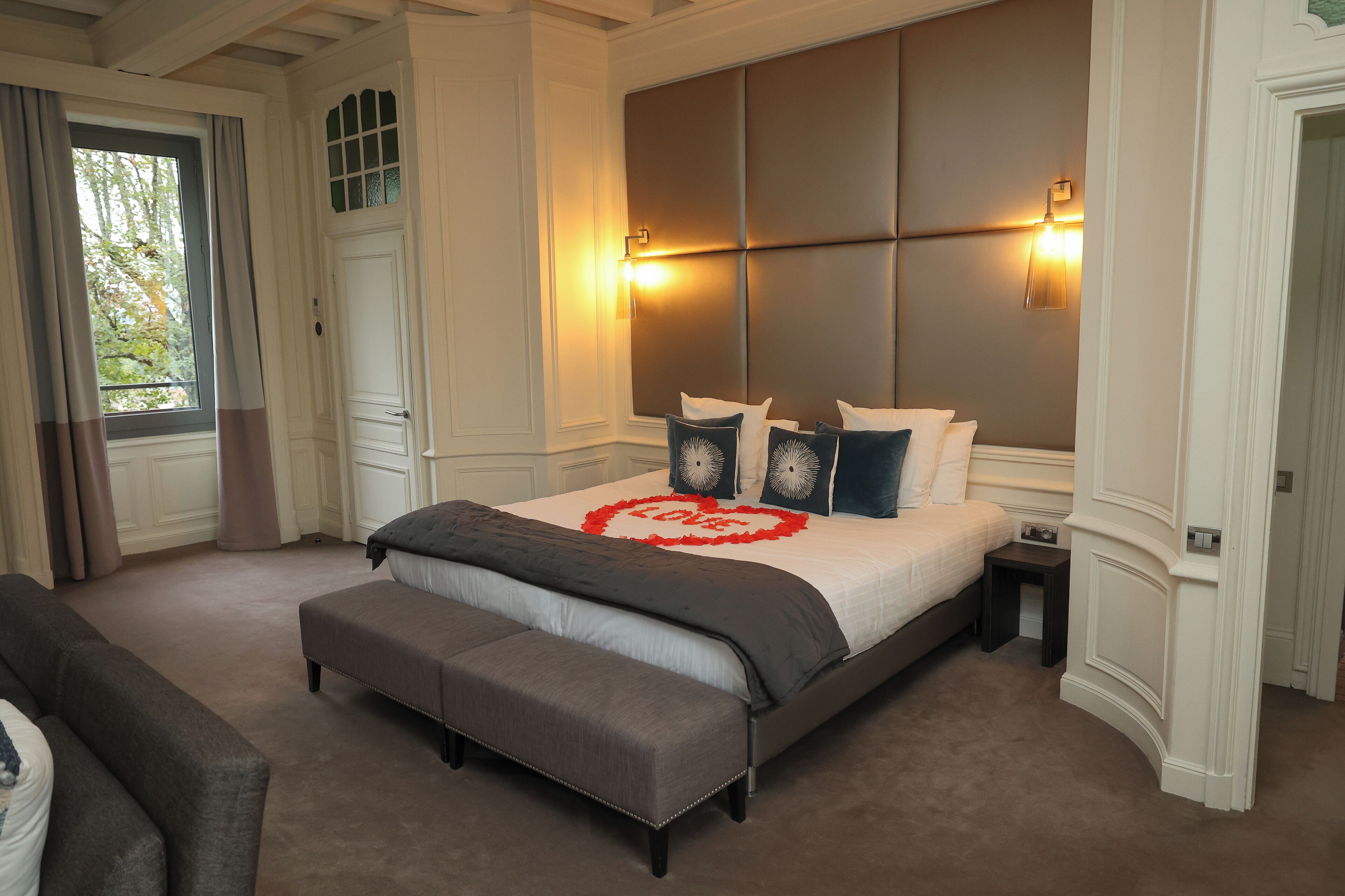 Une chambre avec un grand lit, une banquette, une table de chevet et une fenêtre avec des rideaux.