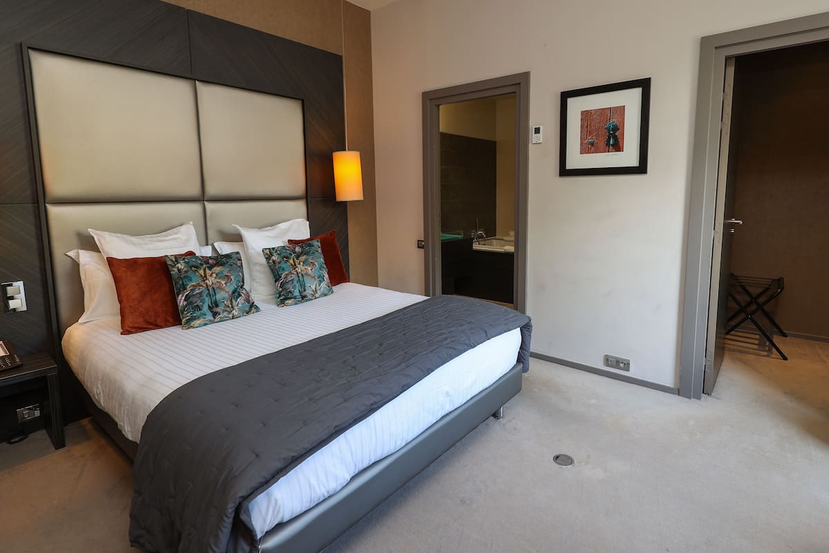 A neatly made bed with a grey bedspread and multiple pillows, a bedside table with a lamp, and a framed picture on the wall.